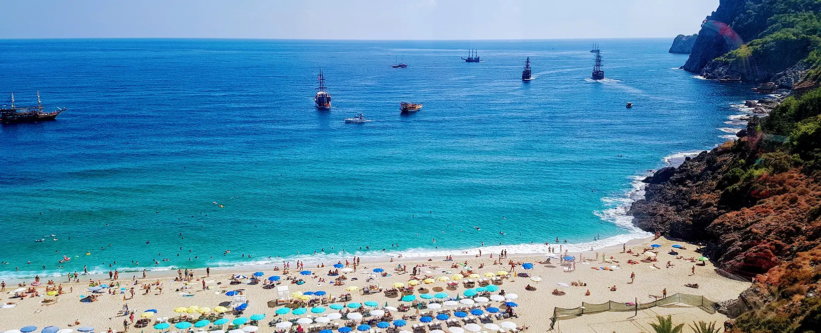 Plaja din Alanya, Turcia, cu nisip auriu, apa azurie a Marii Mediterane cu nave, palmieri, randuri de sezlonguri si umbrele colorate